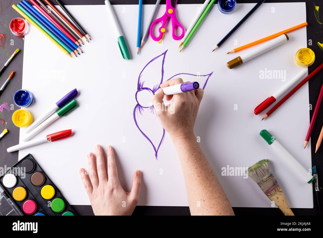 Testa delle mani che disegnano fiori viola su carta, con materiali artistici sul piano del tavolo Foto Stock