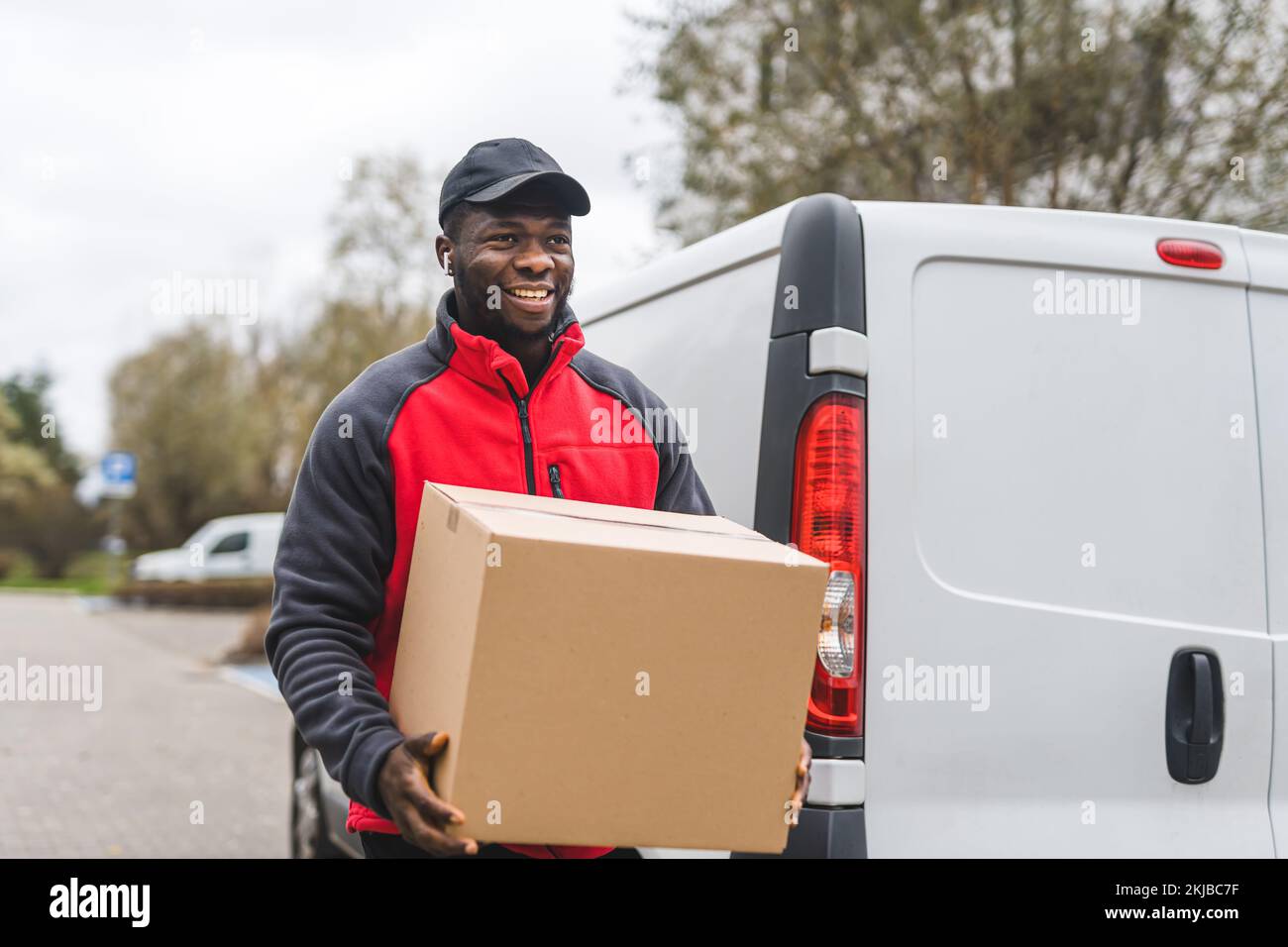 Giovane adulto nero consegna ragazzo indossare uniforme lavoro sorridente trasporto confezione di cartone scaricato da furgone bianco. Scatto orizzontale all'aperto. Foto di alta qualità Foto Stock