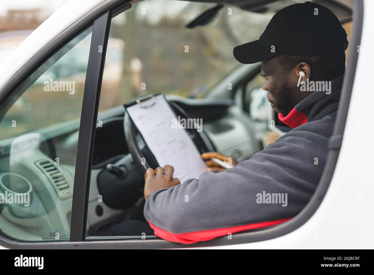 Primo piano di un giovane adulto che indossa un pullover nero e un cappellino nero seduto al posto di guida del furgone bianco che controlla i documenti compilati. Scatto orizzontale. Foto di alta qualità Foto Stock