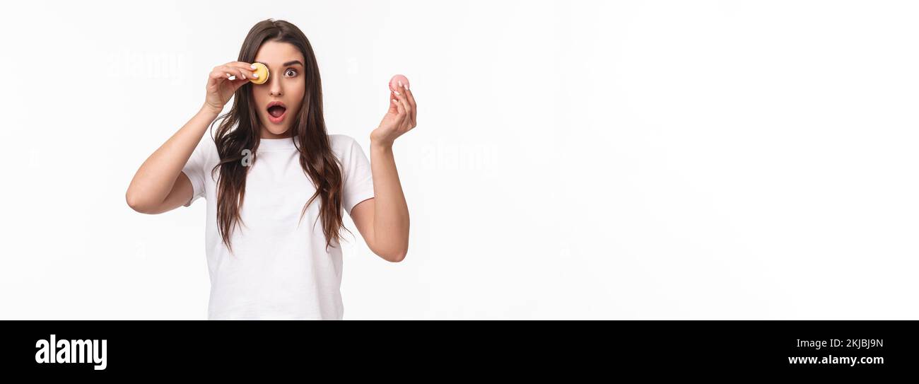 Ritratto di ragazza guardando sorpreso la macchina fotografica con la bocca aperta, guardare qualcosa di sorprendente e impressionante, tenere macaron sopra l'occhio, in piedi bianco Foto Stock