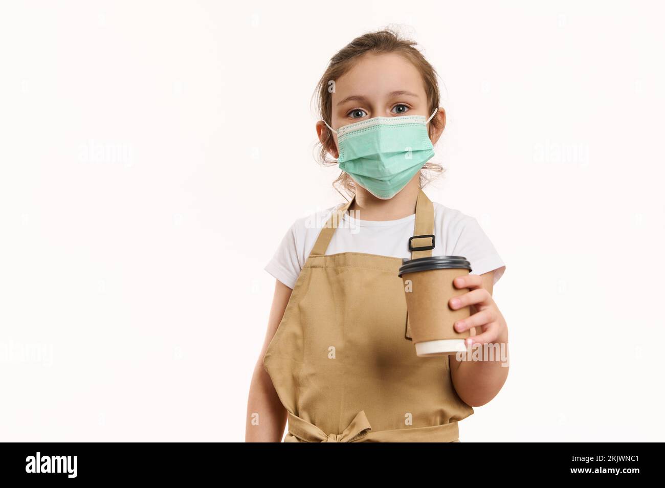Adorabile bambina in grembiule dello chef e maschera medica protettiva verde, distribuendo una bevanda calda da asporto in tazza di carta Foto Stock