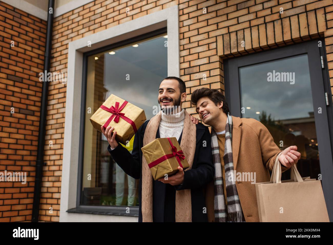 Barbuto gay uomo che tiene le scatole del regalo di Natale vicino al ragazzo allegro e negozio con le vetrine, immagine di scorta Foto Stock
