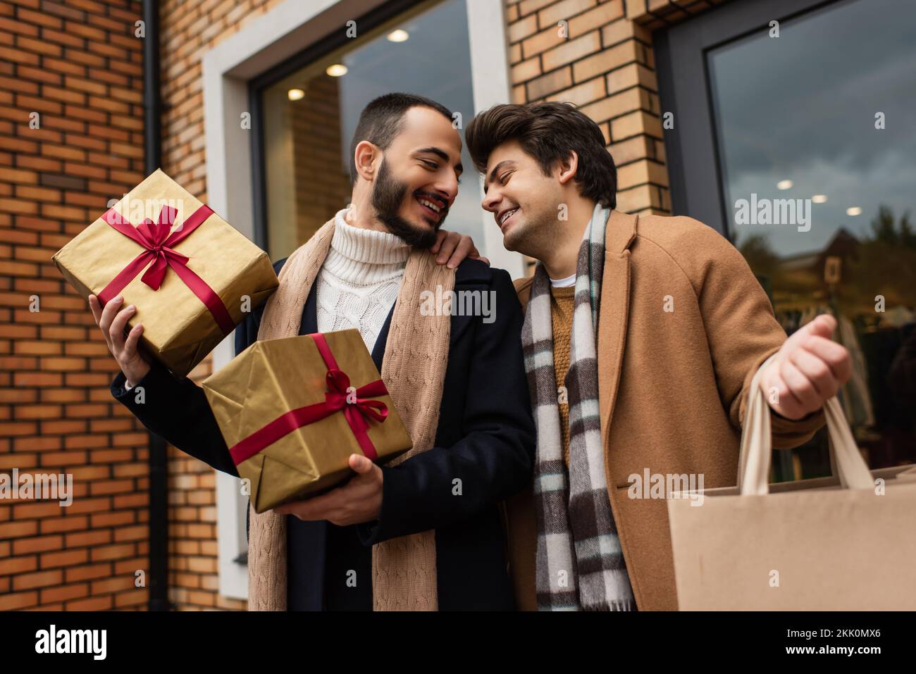 Coppia gay trendy con regali di Natale e shopping bag sorridente vicino al negozio con vetrine sulla strada della città, immagine stock Foto Stock