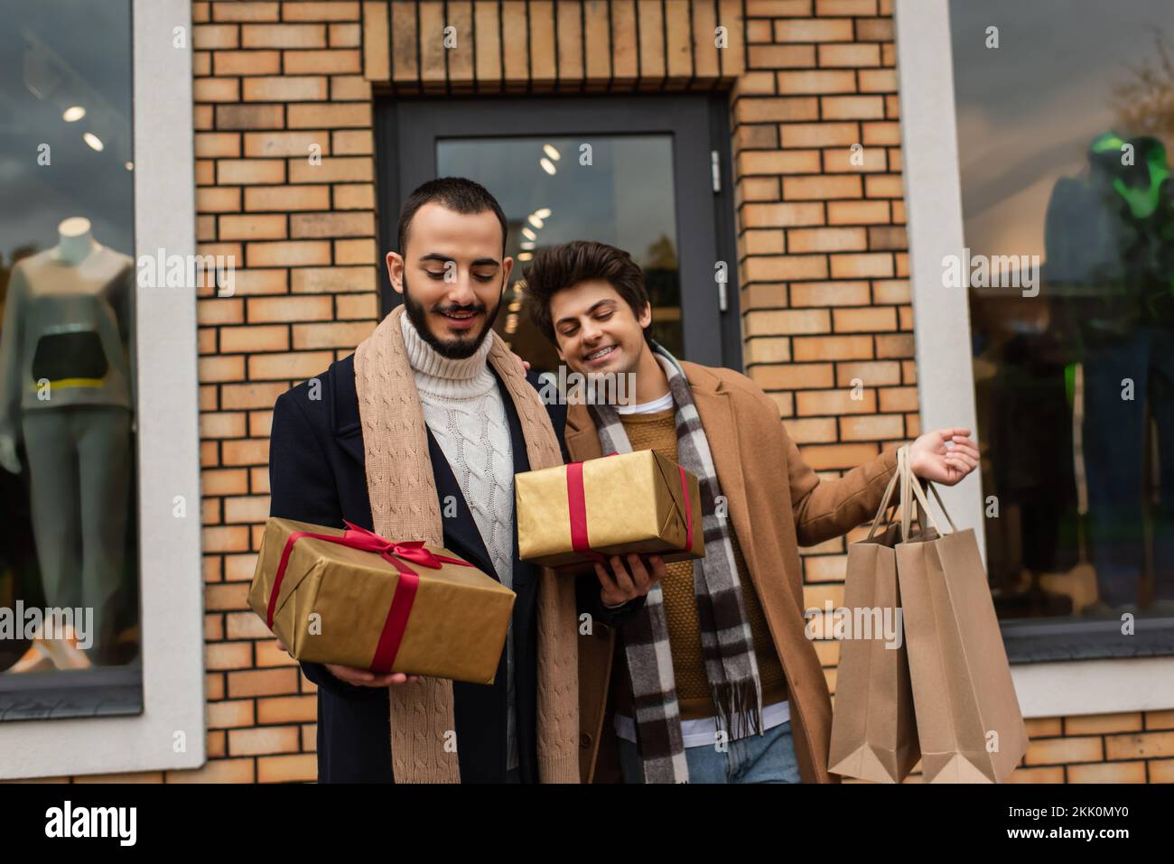 Elegante coppia gay in possesso di scatole regalo di Natale e borse per la spesa vicino al negozio con vetrine sullo sfondo, immagine stock Foto Stock