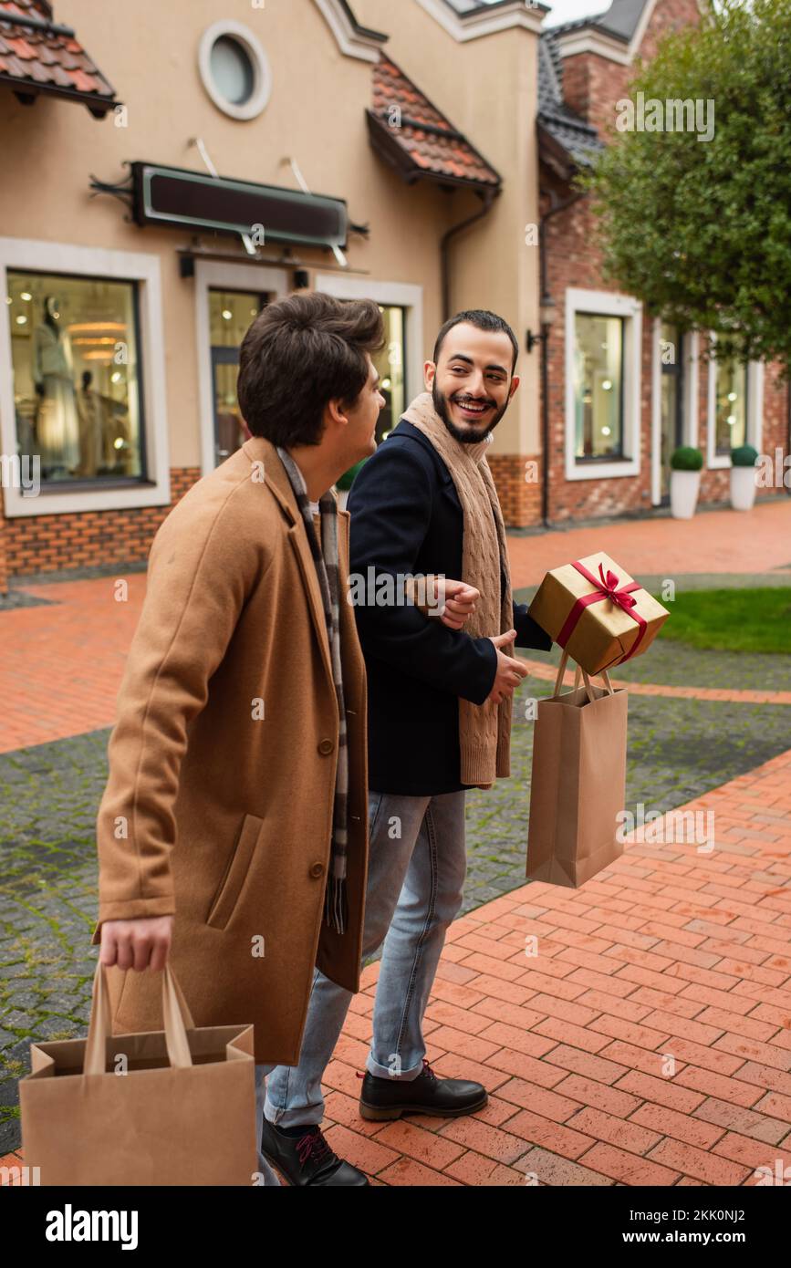 uomini gay felici con borse per lo shopping e scatola regalo a piedi sulla strada con negozi sfocati, immagine stock Foto Stock