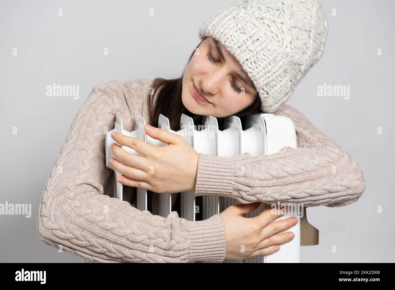 Una donna brunetta in un cappello abbraccia un riscaldatore ad olio, si riscalda in inverno. Foto Stock
