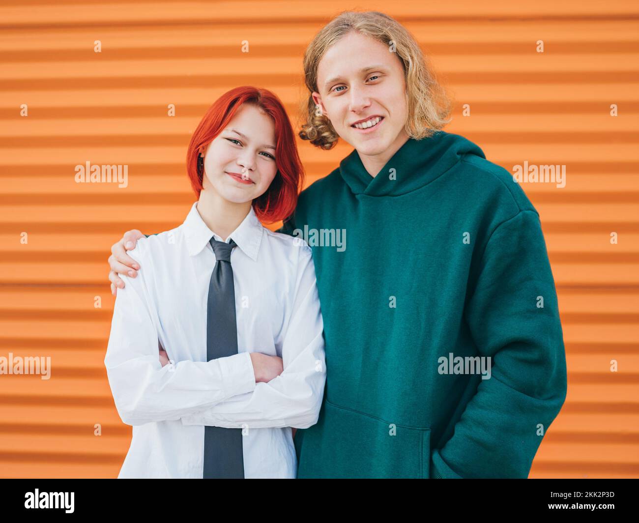 Ritratti di due amici giovani caucasici sorridenti ragazzo a capelli lunghi e bambina di capelli tinti in posa per la foto mentre si trovano vicino alla parete arancione backgroun Foto Stock