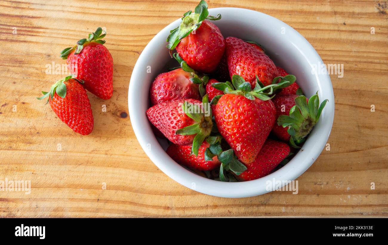 Vista piatta di una ciotola di ceramica bianca con fragole fresche rosse all'interno e due di esse poste sopra un tavolo di legno Foto Stock