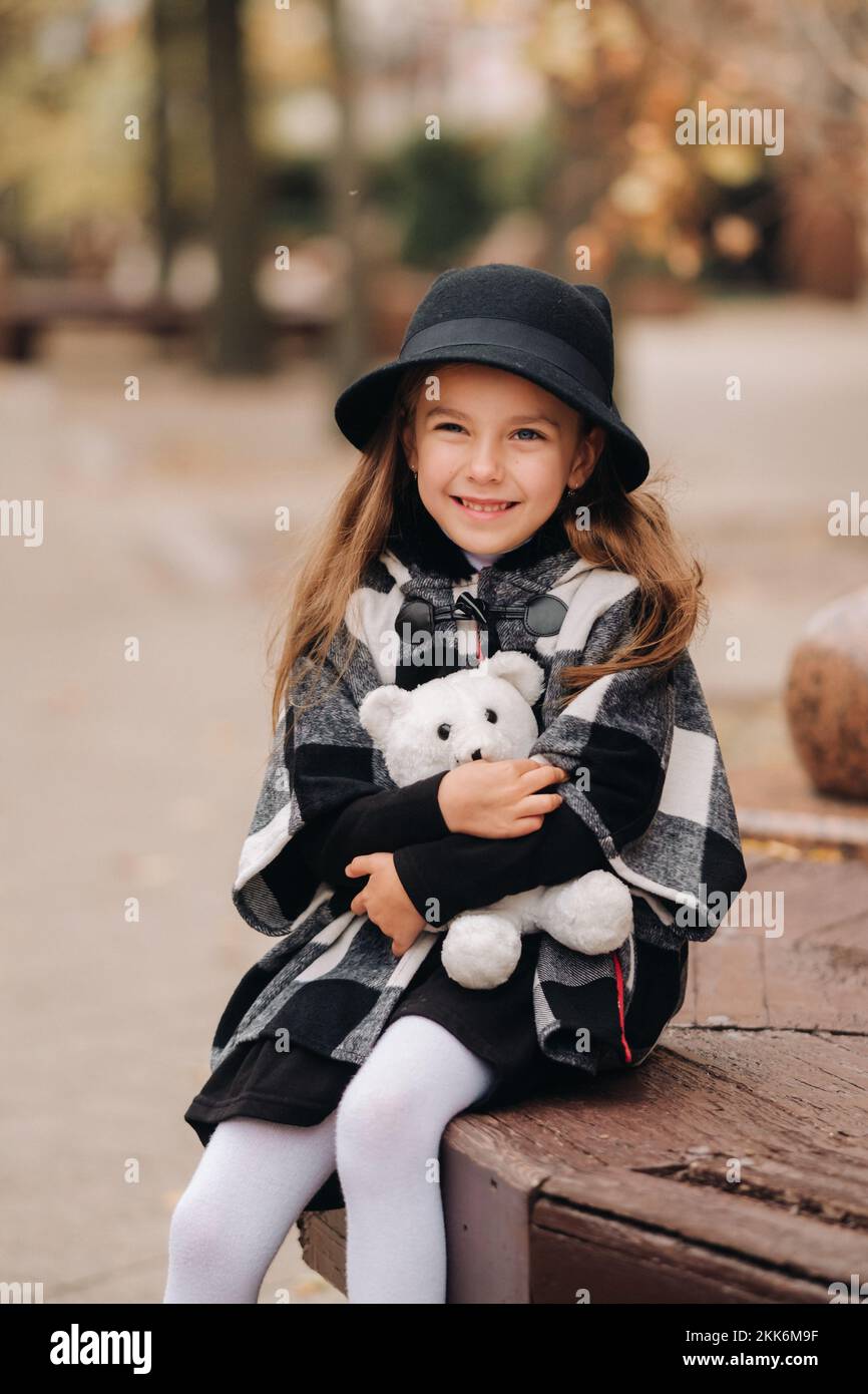 Una bambina piccola ed elegante in abiti autunnali è seduta su una panchina nella città autunnale Foto Stock