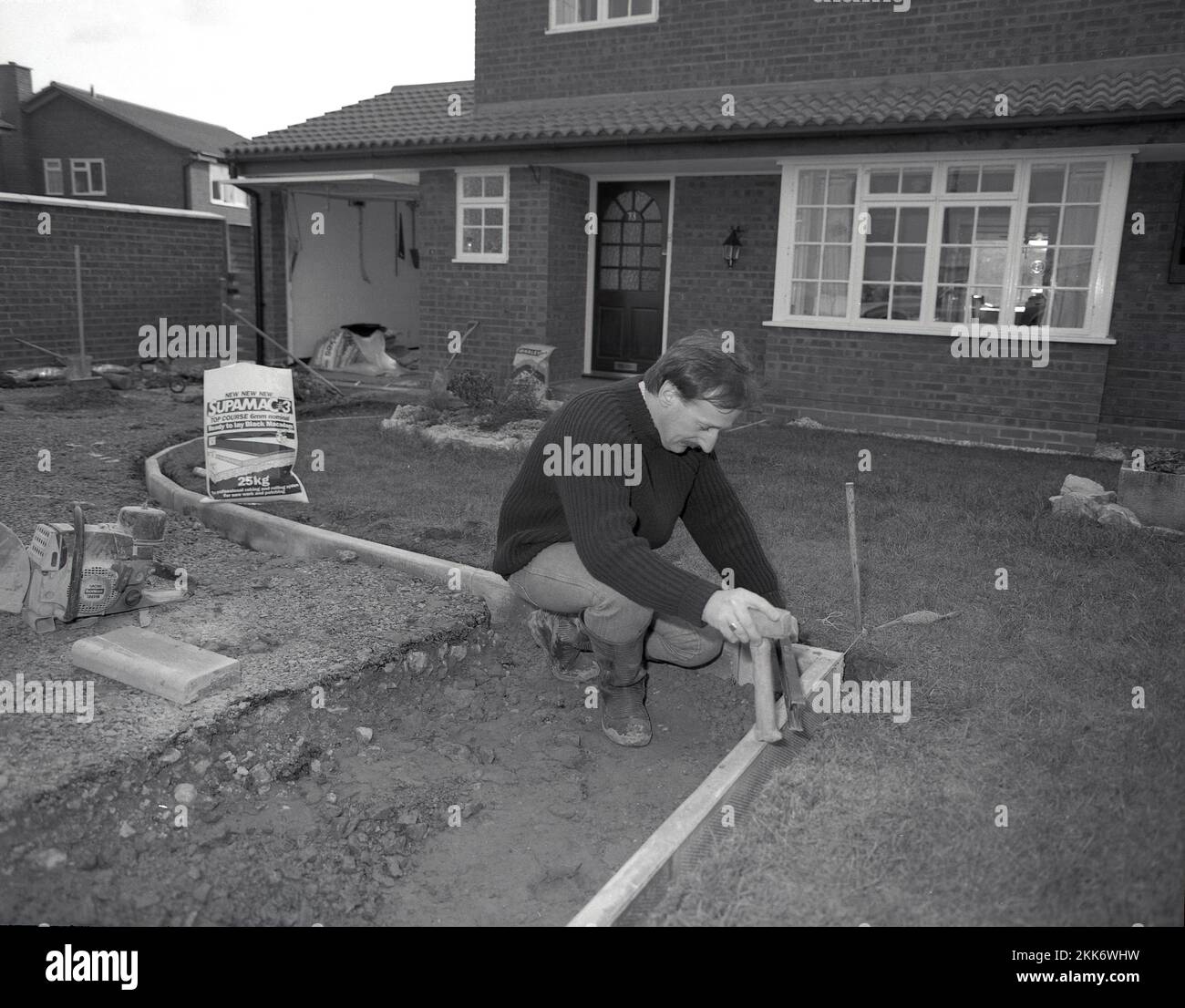 1980s, uomo che costruisce un vialetto, sentiero davanti ad una casa indipendente, che segna il confine concreto, Inghilterra, Regno Unito. usando pronto a signora macadam nero. Foto Stock