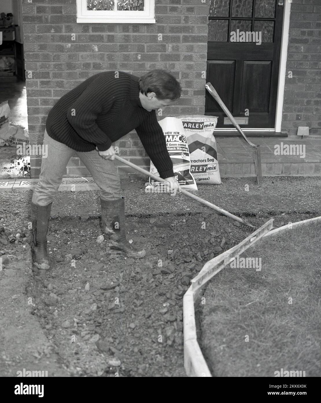 1980s, costruzione, un uomo che estende e riapre un'area su un vialetto e un sentiero davanti ad una casa, utilizzando un rastrello per spargere un corso di aggregato, posato contro nuovi bordi di cemento, Inghilterra, Regno Unito. Foto Stock