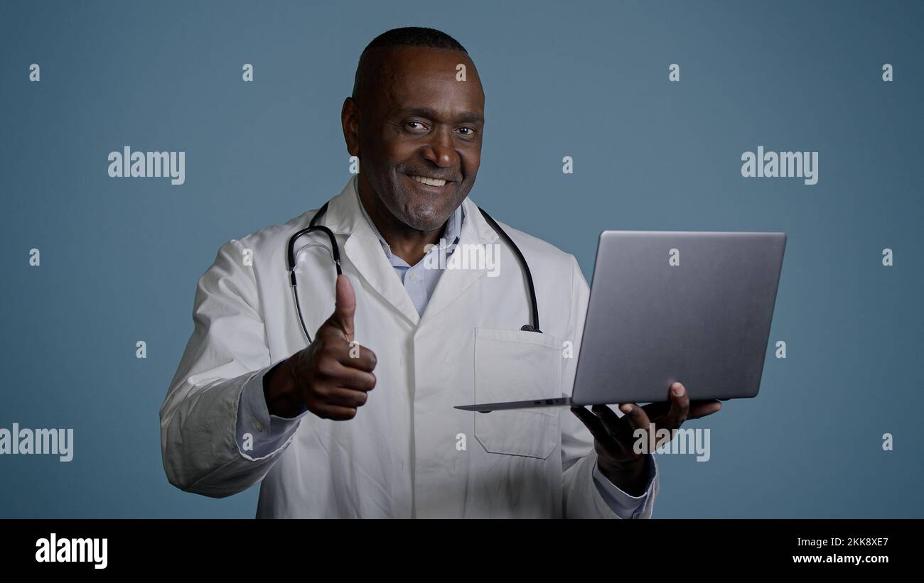 Uomo maturo si alza in studio su sfondo grigio medico professionale in test di controllo abito medico utilizzando laptop consulta paziente online mostrando pollice in alto Foto Stock