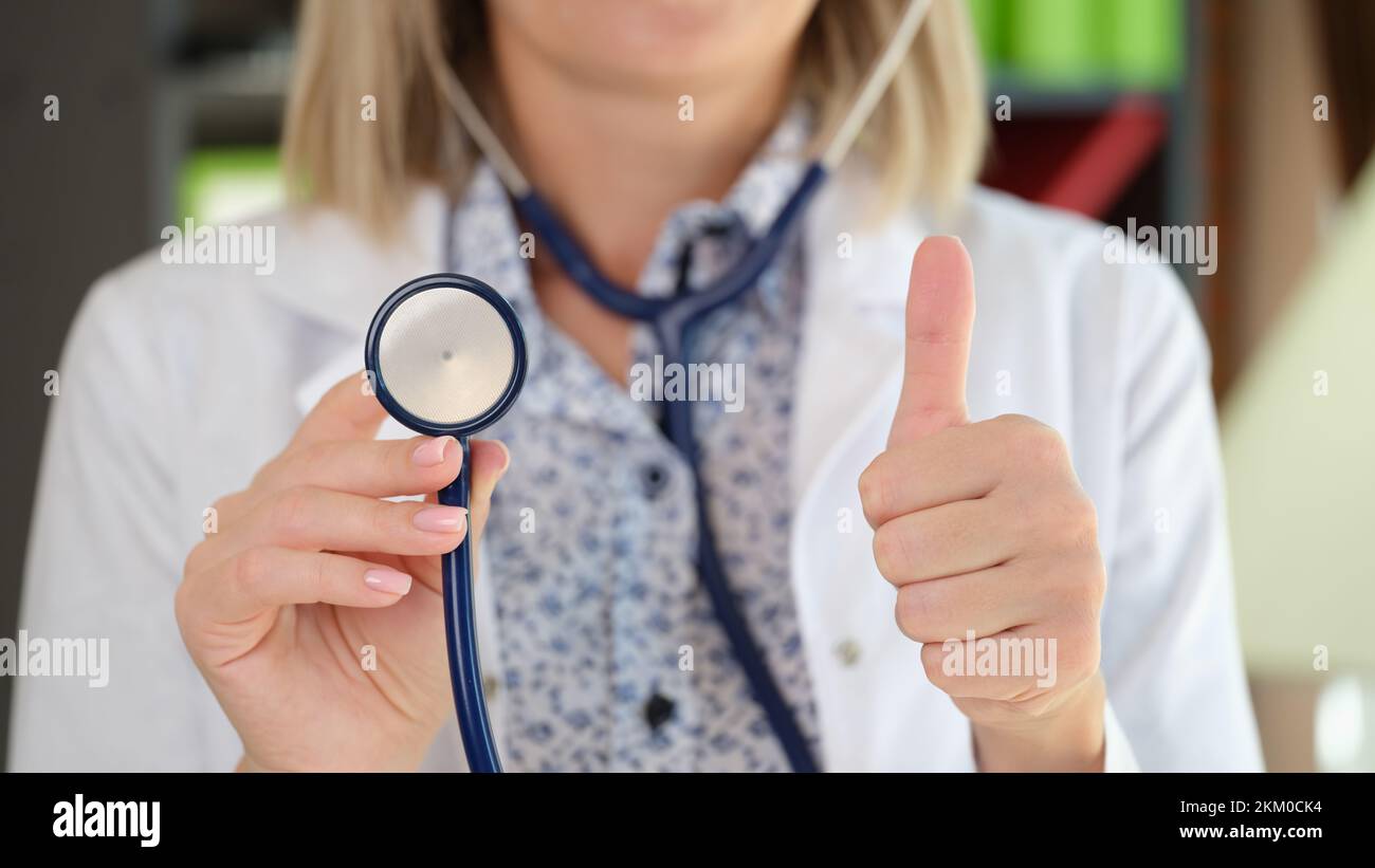 Donna praticante che si pone in clinica ufficio mostrando stetoscopio e pollice verso l'alto gesto Foto Stock