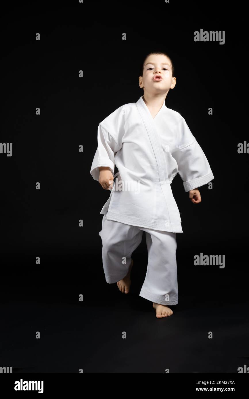 Il ragazzino forte in kimono va avanti, pratica il karate, sfondo nero Foto Stock