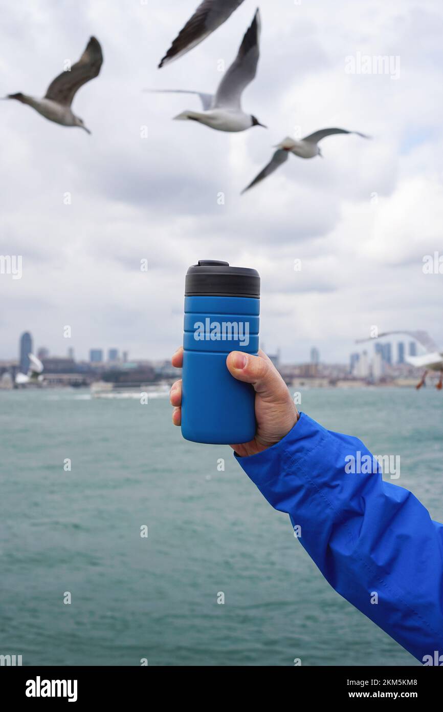 Mano maschile che tiene una tazza di caffè da viaggio thermos sullo sfondo di acqua limpida di un mare con una tonalità turchese. Foto Stock