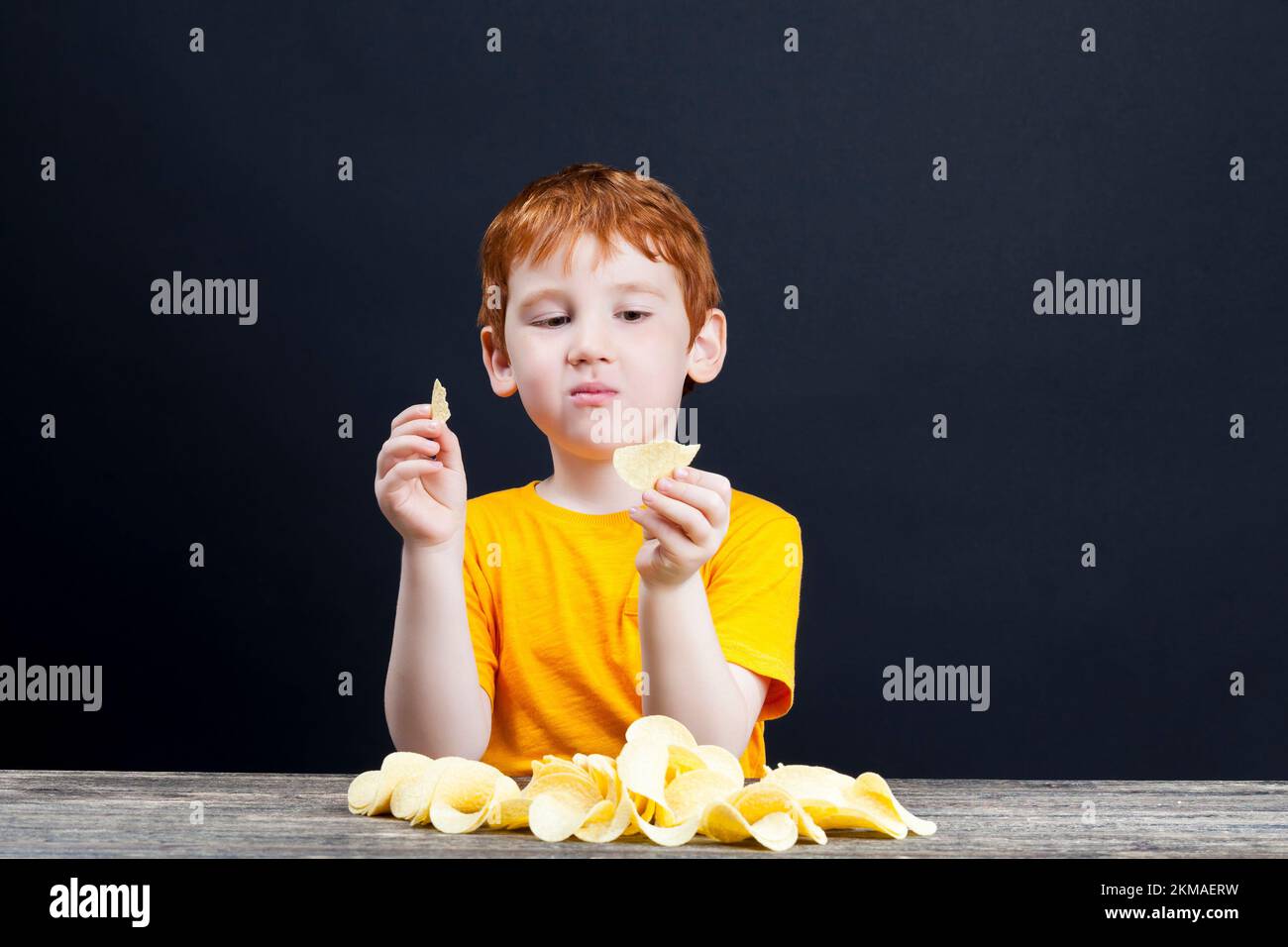 patatine croccanti deliziose che un bambino con i capelli rossi mangia, cibo nocivo, ma che il bambino vuole davvero mangiare, il ragazzo al tavolo, vicino Foto Stock