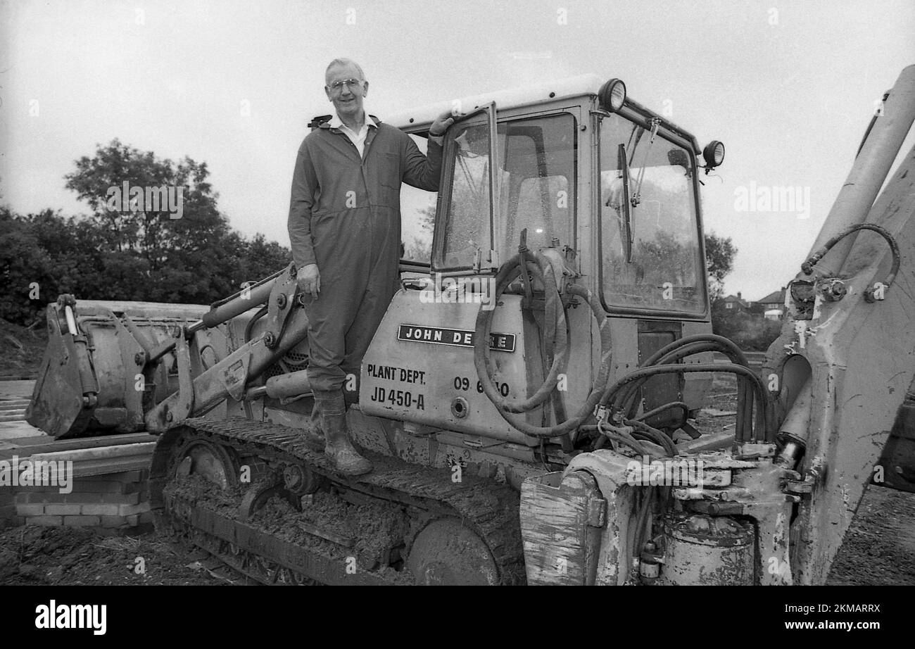 1980s, storico, all'esterno di un cantiere edile, un costruttore in tute monopezzo su una pala cingolata John Deere 450, Inghilterra, Regno Unito. Il modello 450 è stato introdotto per la prima volta nel 1965 ed è stato un popolare bulldozer per molti anni. Foto Stock