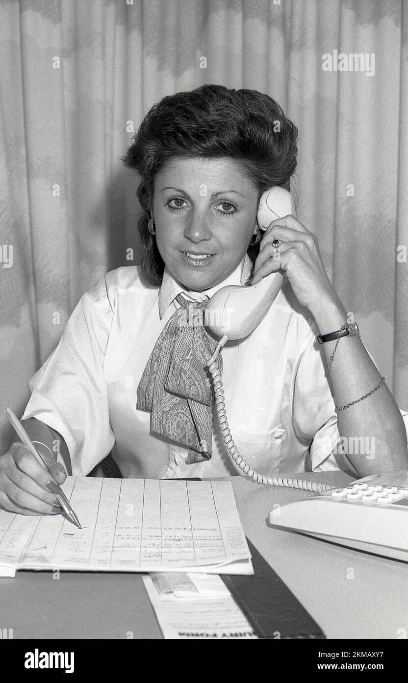 1980s, storica, una donna di vendita di nuove case che indossa una blusa, con colletto e sciarpa di seta alla scrivania al telefono, con un calendario di appuntamenti su un foglio di carta, Inghilterra, Regno Unito. Foto Stock