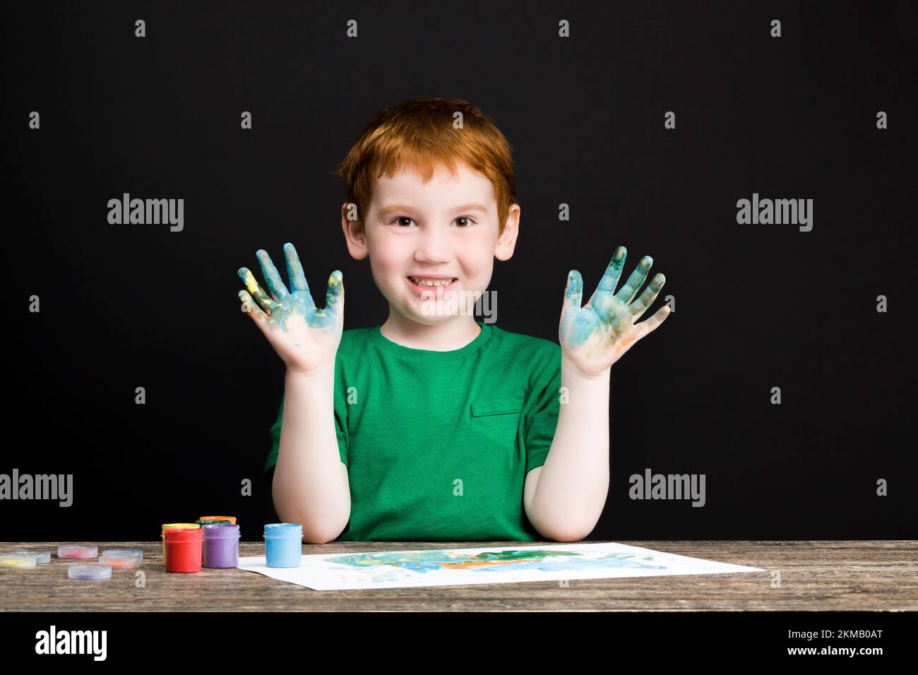 ritratto di un ragazzo felice dai capelli rossi attinge su carta utilizzando vernici di colori diversi, il ragazzo si disegna con le mani e così le sue mani sono dipinte Foto Stock