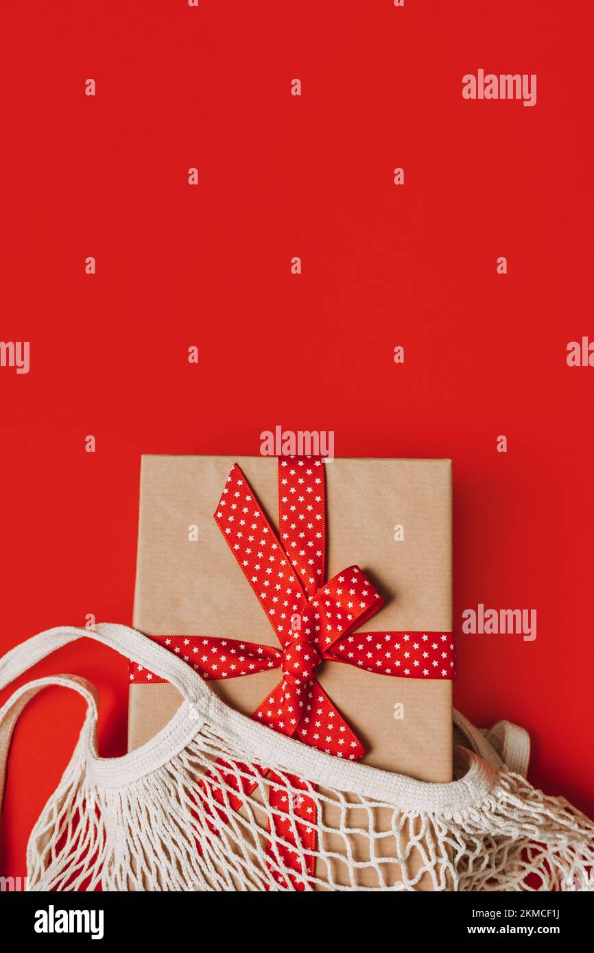 Natale eco friendly zero rifiuti idee regalo. Xmas moderno cotone Mesh Grocery net bag con scatola regalo in carta kraft nastro rosso su sfondo rosso. Foto Stock