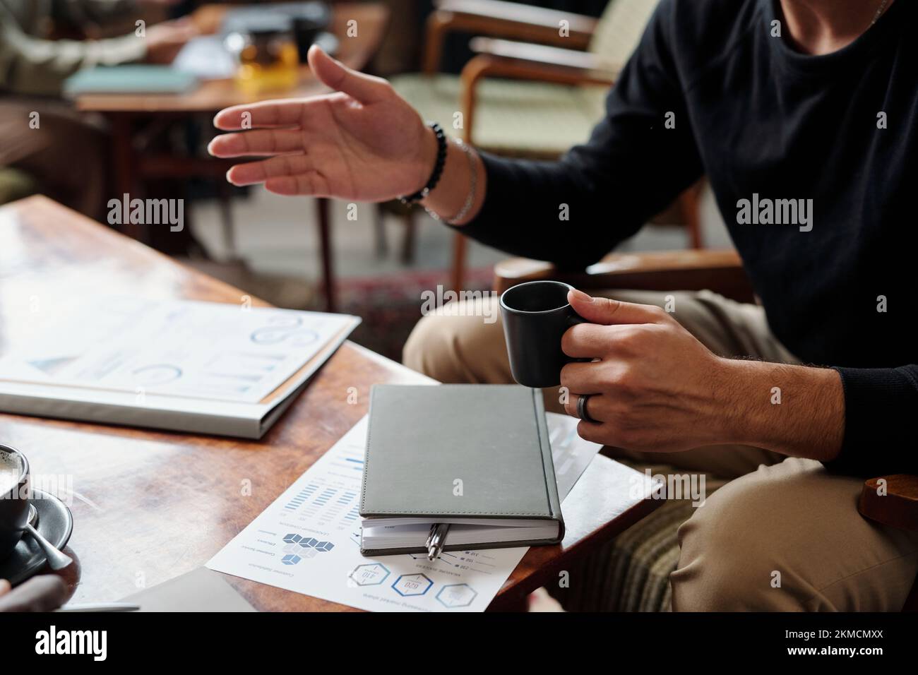 Mani di un giovane economista maschile con una tazza di tè seduto al tavolo con notebook e documenti finanziari durante la conversazione con un collega Foto Stock