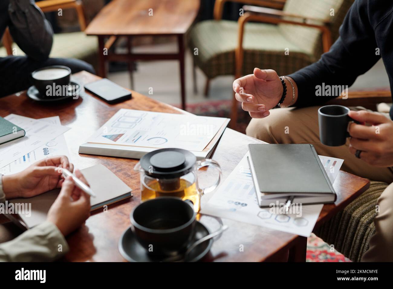 Mani di un giovane uomo d'affari con una tazza di tè seduto al tavolo con documenti finanziari e parlando con i colleghi alla riunione Foto Stock