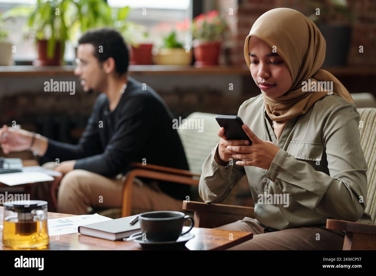Giovane donna d'affari musulmana nella messaggistica hijab in smartphone o organizzando il lavoro mentre si siede al tavolo con tè e notebook in caffetteria Foto Stock