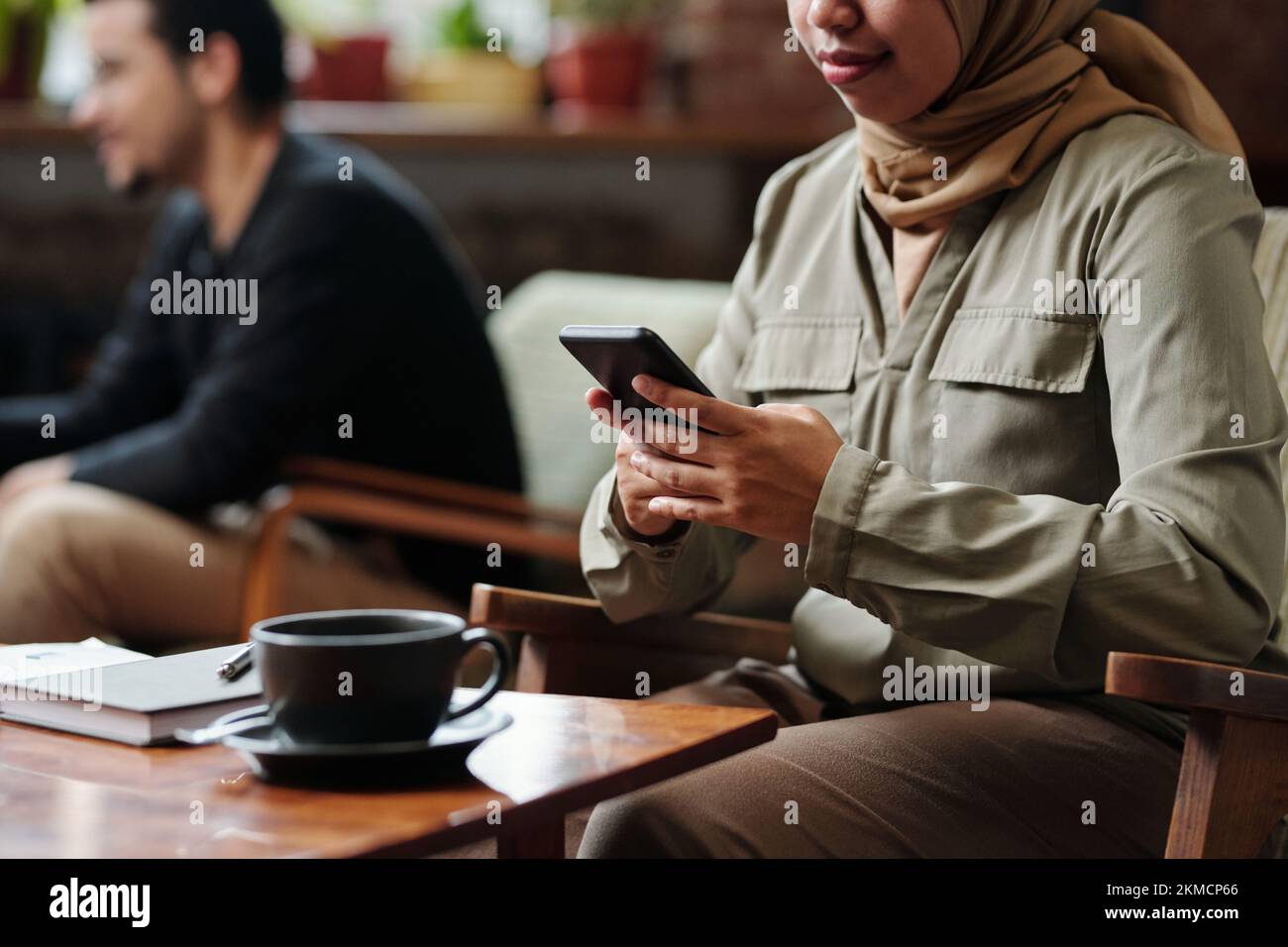 Mani di una giovane dipendente musulmana che utilizza lo smartphone mentre si siede al tavolo con una tazza di tè e un notebook e organizza il lavoro Foto Stock