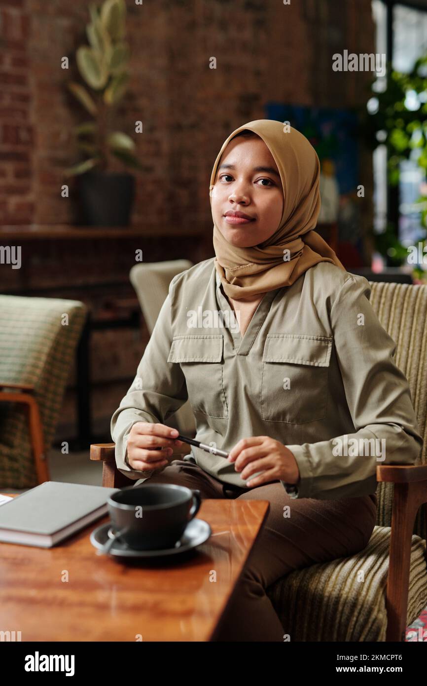 Giovane donna musulmana afro-americana di successo o intern in hijab e casualwear seduti in poltrona e organizzando il lavoro Foto Stock
