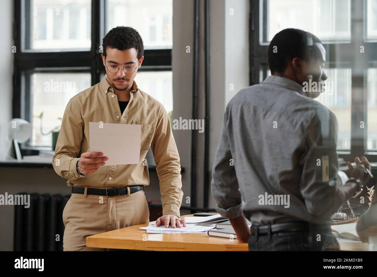 Giovane uomo d'affari serio che guarda attraverso il documento finanziario mentre si trova al tavolo di fronte a un collega afro-americano Foto Stock