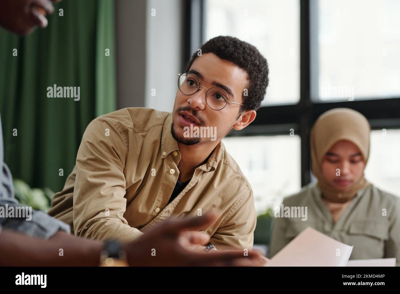 Giovane uomo d'affari multietnico in casualwear e occhiali che parla con un collega afro-americano maschio dopo aver presentato un rapporto finanziario Foto Stock