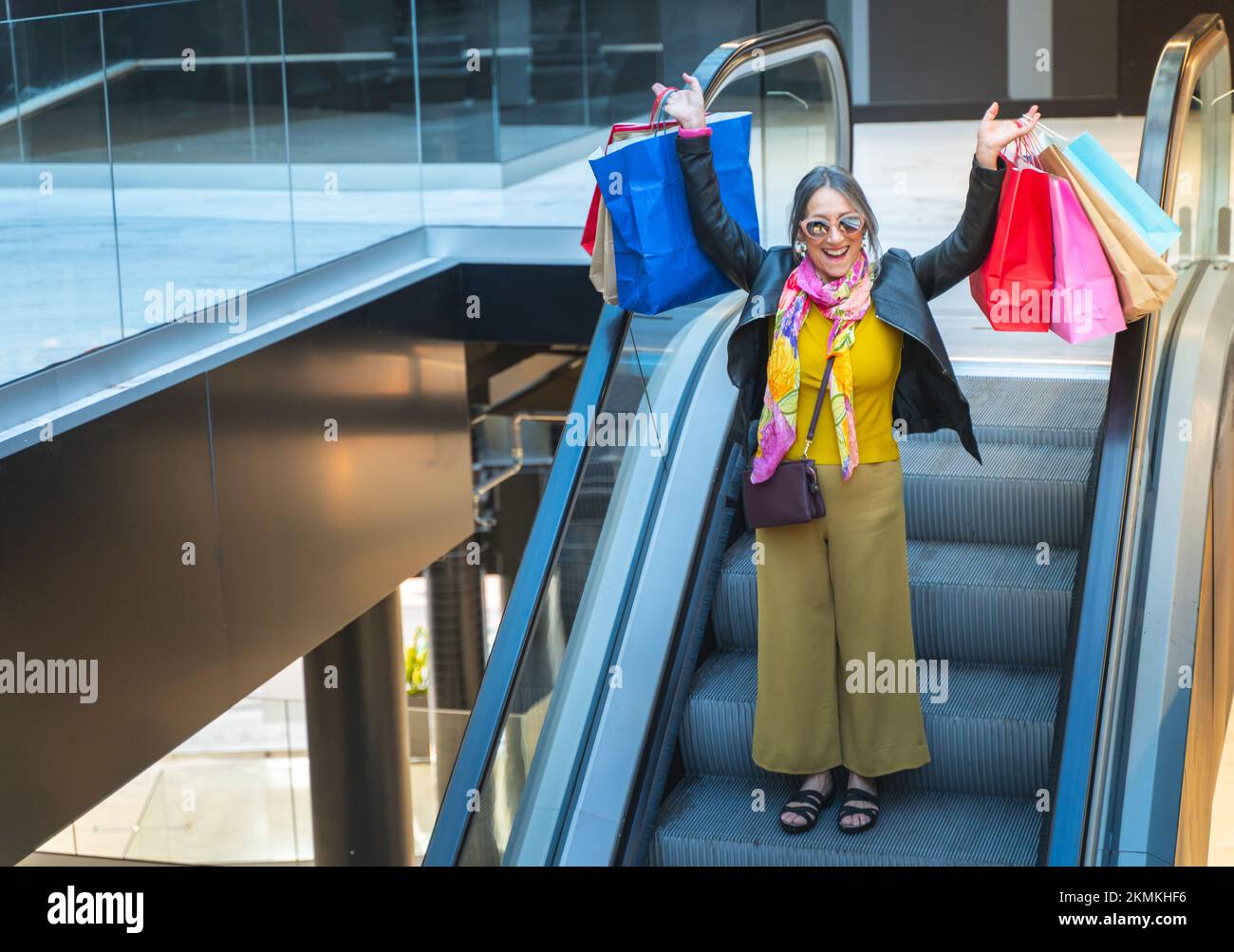 Signora anziana festeggia con le borse della spesa sulle scale meccaniche del centro commerciale. Donna che scende scala mobile. Foto Stock