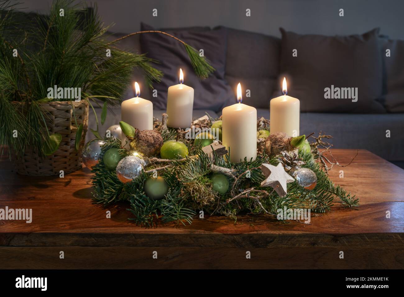Corona dell'Avvento con quattro candele bianche brucianti e decorazione di Natale su una tavola di legno di fronte al divano, decorazione domestica di festa, spazio della copia, seleziona Foto Stock