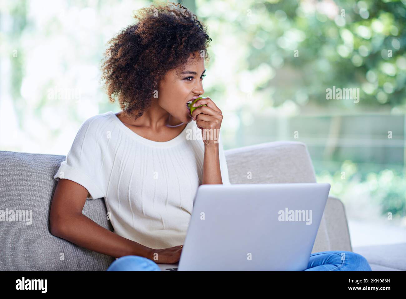 Giornata rilassata a casa con spuntini e tecnologia. una giovane donna che mangia una mela mentre usa il suo notebook a casa. Foto Stock