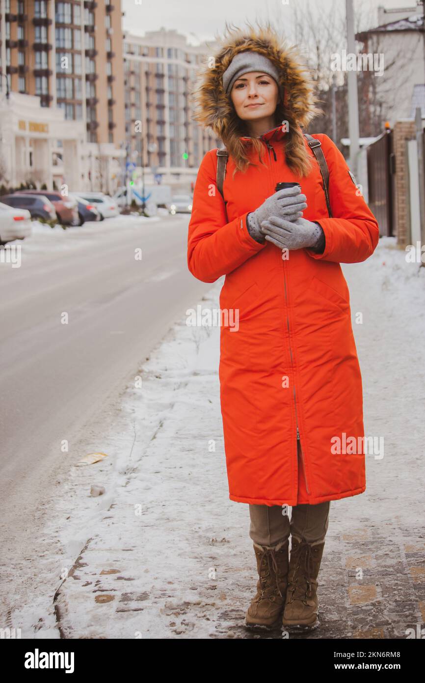 Ragazza con caffè in giacca calda con cappuccio in pelliccia per la strada. Moda invernale. Bella donna in giacca arancione con cappuccio in città. Stile di vita urbano Foto Stock