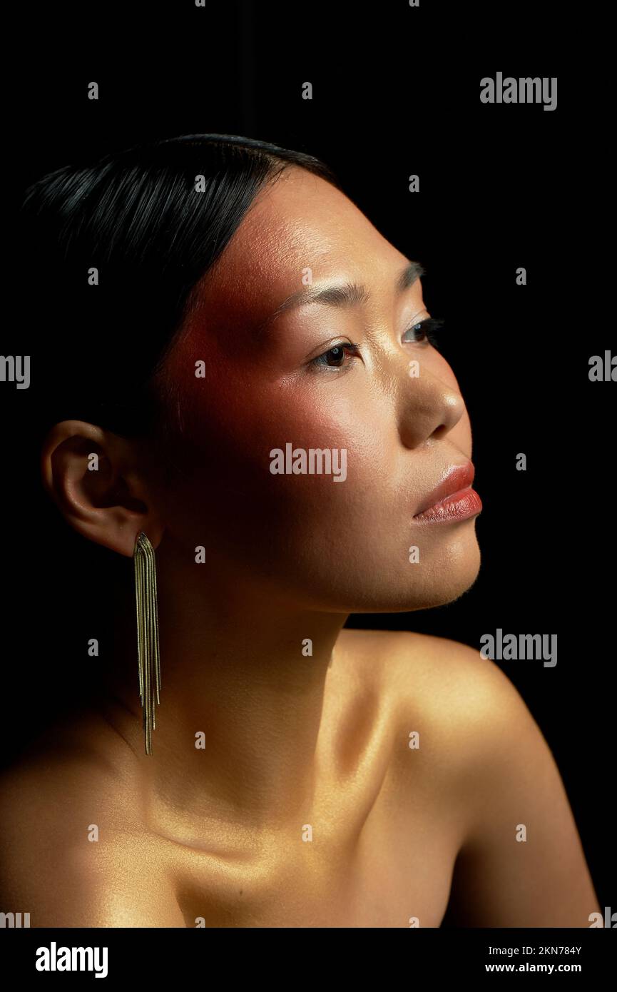 Una ragazza asiatica con pelle dorata guarda nel profilo. Orecchini lunghi in oro. Salone di bellezza. Foto Stock