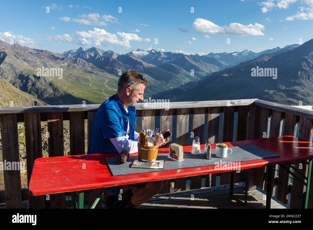 Un escursionista si siede sulla terrazza della capanna montana Brunnenkogelhaus e guarda al suo cellulare di fronte alle Alpi di Ötztal e alla valle di Gurgl, Austria Foto Stock