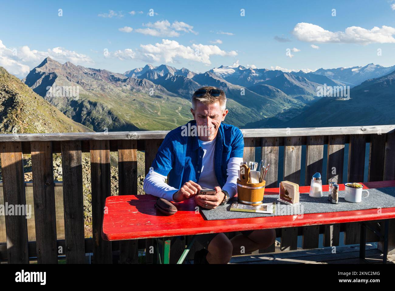 Un escursionista si siede sulla terrazza della capanna montana Brunnenkogelhaus e guarda al suo cellulare di fronte alle Alpi di Ötztal e alla valle di Gurgl, Austria Foto Stock