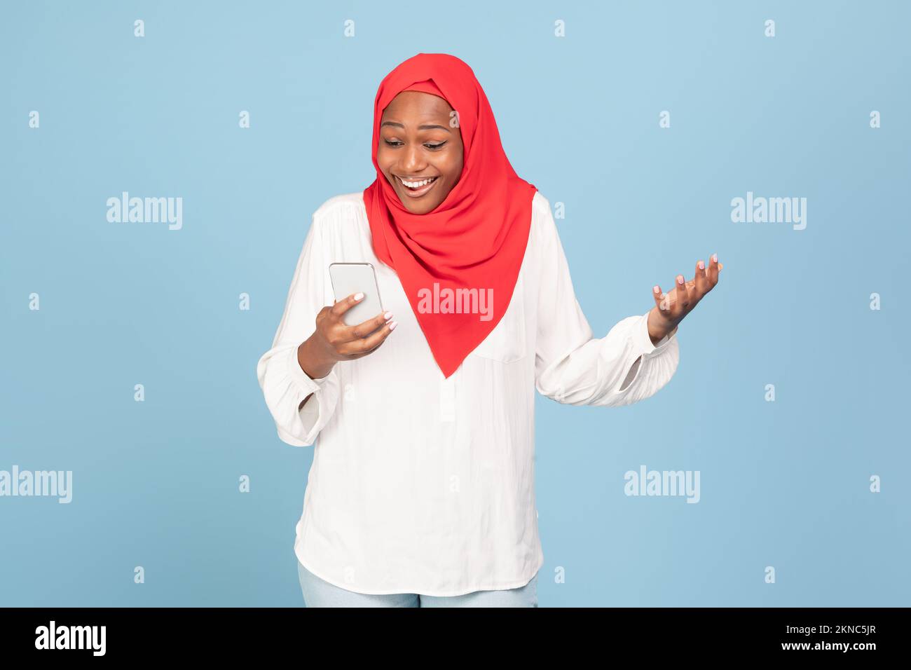 Donna musulmana afro-americana overjoyed che guarda smartphone in eccitazione, posa su sfondo blu studio Foto Stock