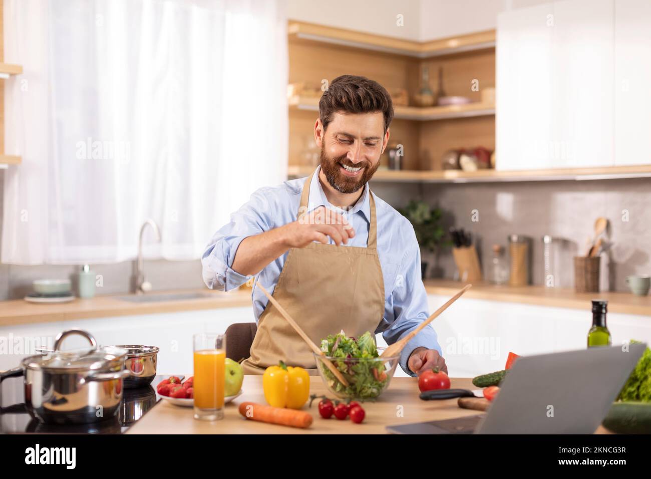 Felice adulto maschio caucasico con barba in grembiule ha videochiamata, facendo video per il blog pasto, insalata salante Foto Stock
