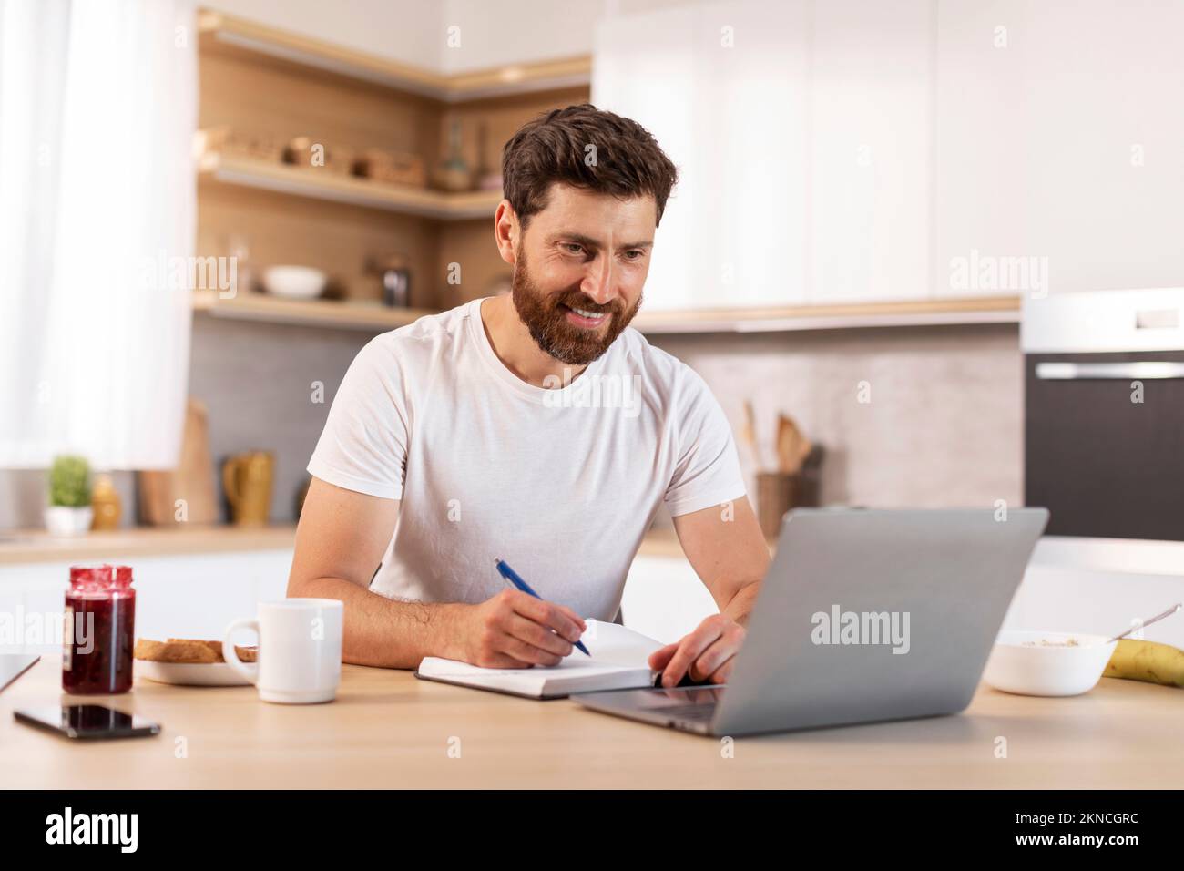 Felice uomo caucasico di mezza età con barba in t-shirt bianca guardare la lezione on-line sul portatile in cucina minimalista Foto Stock