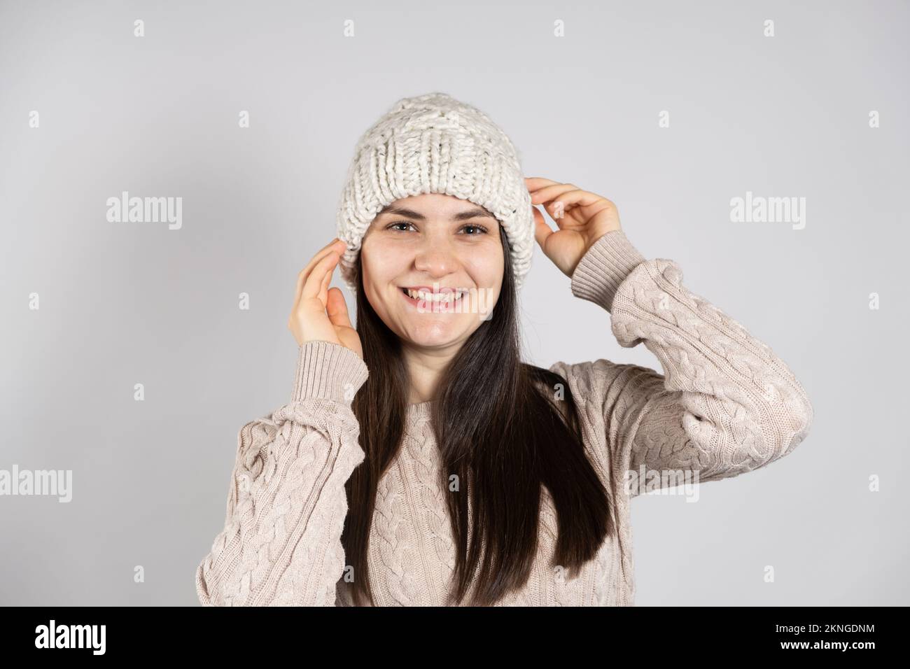 Una donna felice brunetta in un cappello invernale e un maglione lavorato a maglia sorride su uno sfondo bianco. Foto Stock
