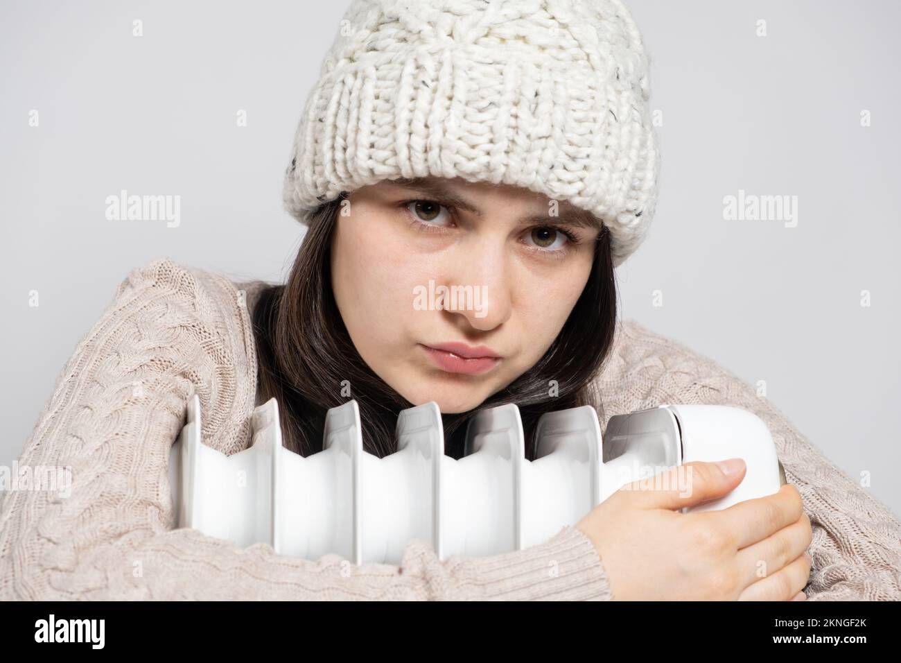 Una donna in un cappello di lana d'inverno abbraccia un riscaldatore a olio elettrico e sembra tristemente, congelato. Foto Stock