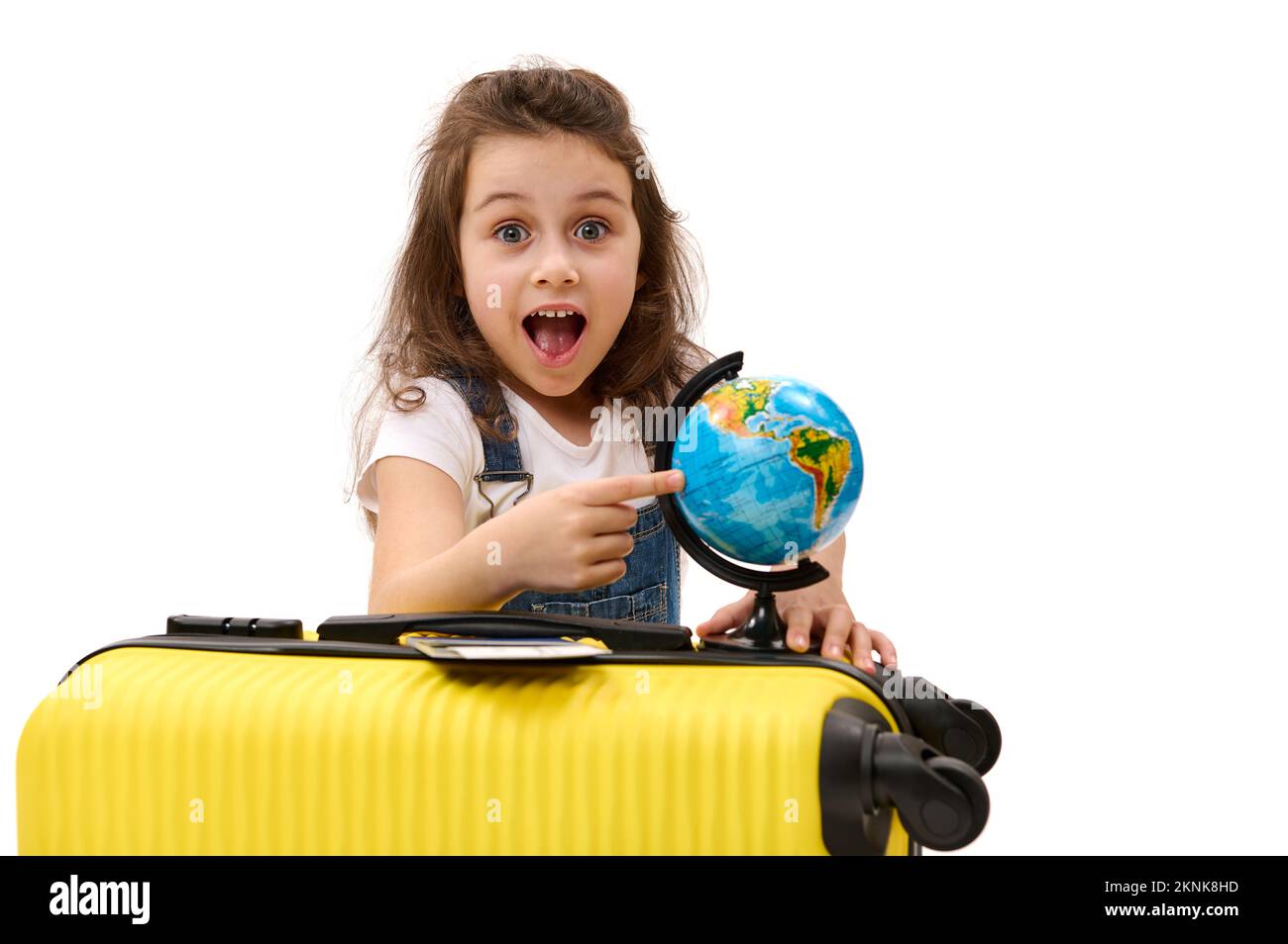 Carina ragazza viaggiatore con una valigia gialla punta a una destinazione sul globo, guardando la macchina fotografica in sorpresa Foto Stock