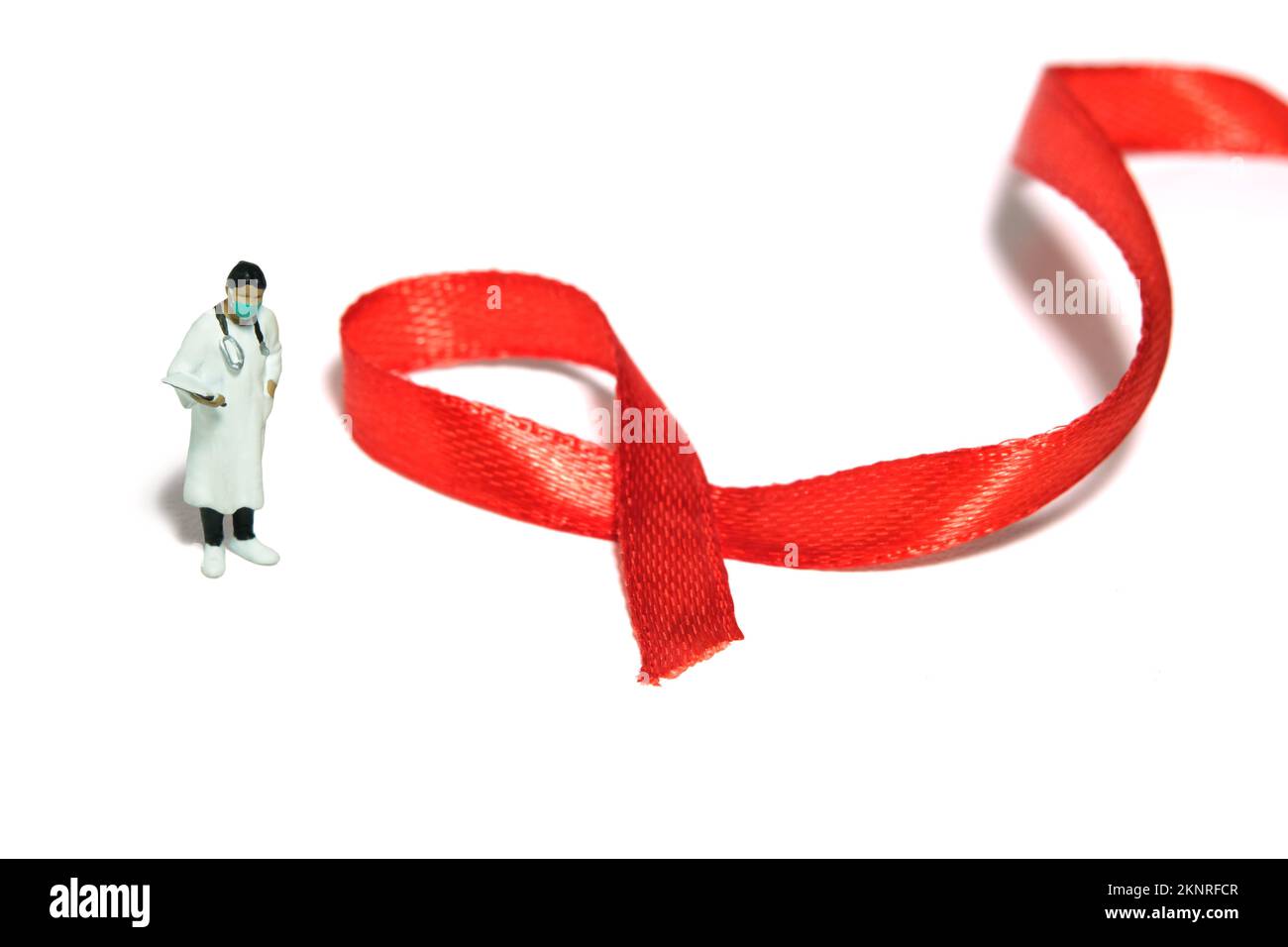 Fotografia di giocattoli di figura della gente in miniatura. Giornata mondiale dell'AIDS concetto di giornata di consapevolezza. Ragazza donna medico in piedi accanto al nastro rosso. Isolato su fondo bianco gr Foto Stock