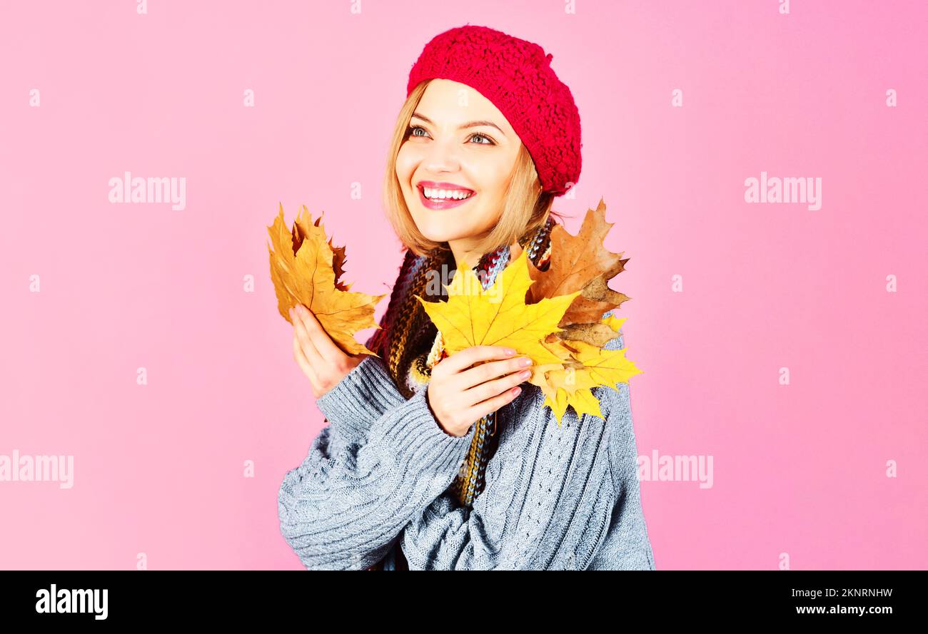 Donna sorridente in abiti caldi con foglie gialle. Ragazza autunnale in cappello rosso, maglione a maglia e sciarpa. Foto Stock
