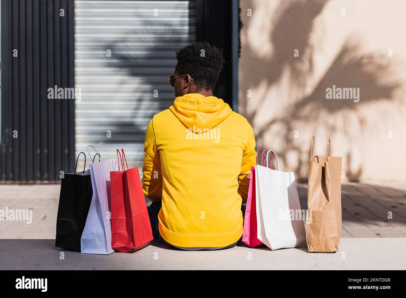Un giovane uomo africano vestito con una felpa gialla seduta su una panca di strada accanto ad alcune borse della spesa visto dalla sua schiena. Foto Stock