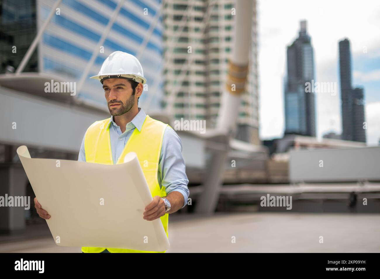 Uomo ingegnere caucasico, indossando un giubbotto giallo e un grande elmetto, e tenendo la mano la pianta bianca del pavimento nel lavoro sito del centro città. Foto Stock