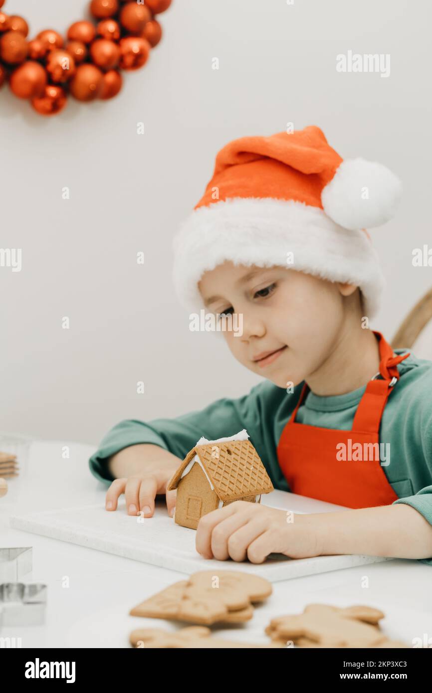 Un ragazzo carino la vigilia di Capodanno e Natale fa una casa di zenzero in cucina in un cappello di Babbo Natale. Foto Stock