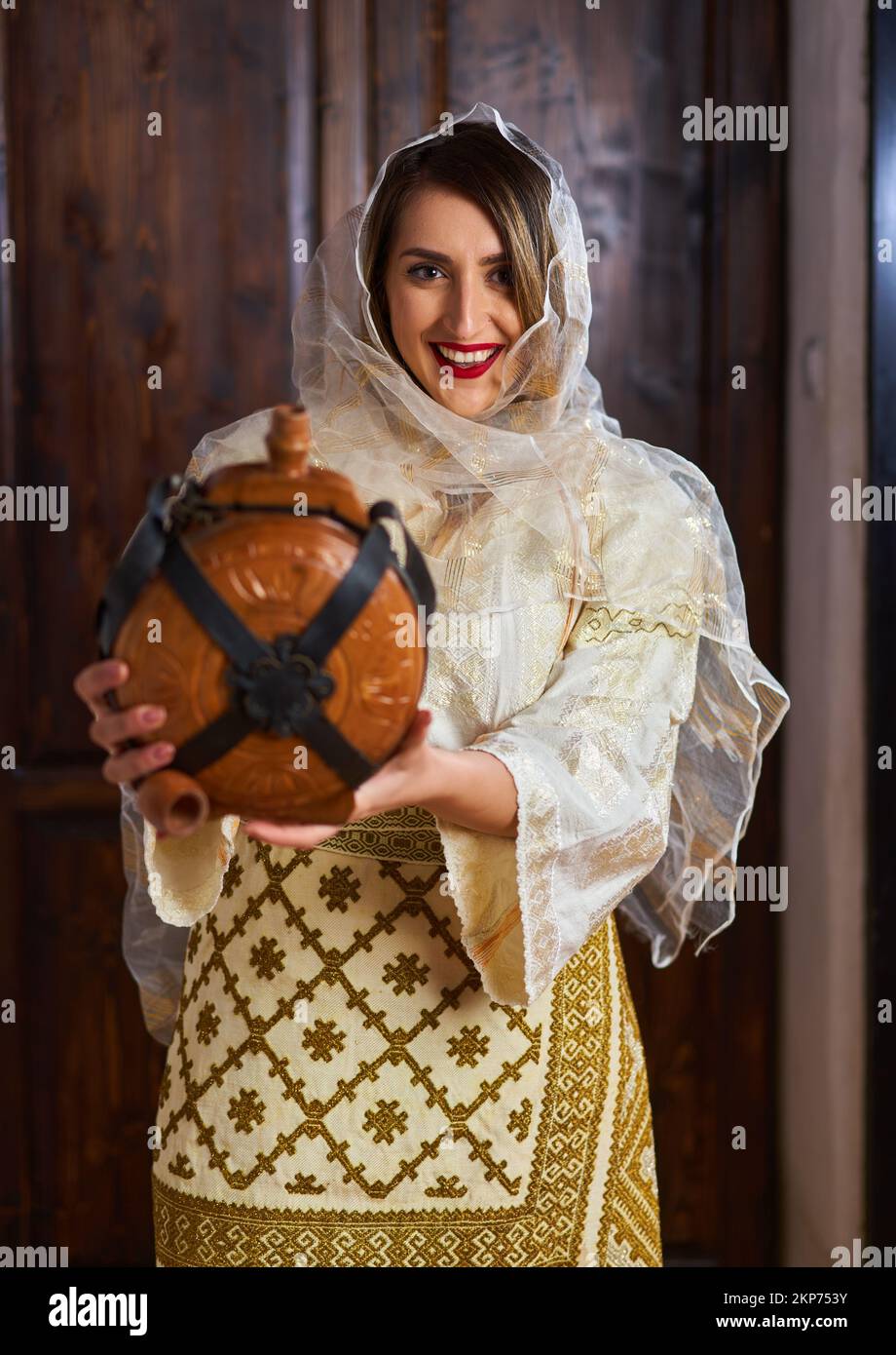 Giovane donna rumena in costume tradizionale sposa popolare in una casa d'epoca con una bottiglia di vino in legno Foto Stock